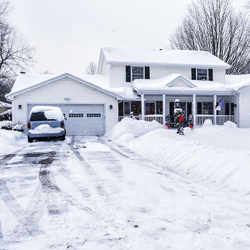 Streetside view of a Christmas holiday decorated residential district single family house front facade during a winter snow storm with snowflakes blowing everywhere. The driveway was recently plowed clear of snow, but is already covered again. Shot taken from the street at the end of the driveway across piled snowdrifts and deep snow on the front yard.