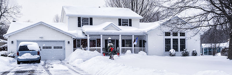 Streetside view of a Christmas holiday decorated residential district single family house front facade during a winter snow storm with snowflakes blowing everywhere. The driveway was recently plowed clear of snow, but is already covered again. Shot taken from the street at the end of the driveway across piled snowdrifts and deep snow on the front yard.